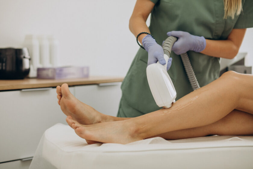 Clinician in green scrubs gliding a white handheld device over a patient’s leg during a cosmetic treatment.