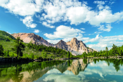 Mountain landscape with green hills, rocky peaks, and a calm lake reflecting the blue sky and white clouds.