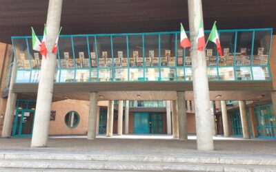 Exterior view of a brick building with a blue railing balcony displaying posters, supported by concrete pillars; multiple Italian flags hang from the railing and columns.