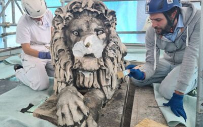 Two workers in helmets and gloves restore a weathered stone lion statue on a scaffolded platform.