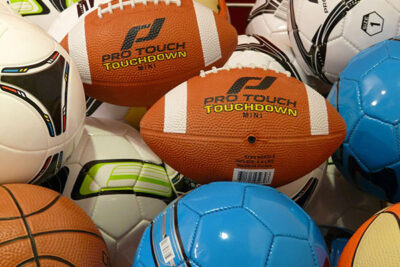 Pile of assorted sports balls in bright colors, including orange footballs, white soccer balls, and blue balls.
