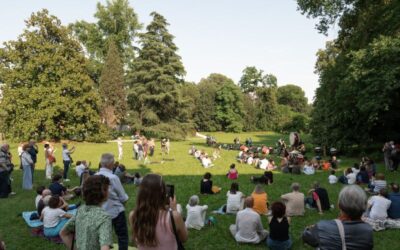 Crowd gathered in a sunny park lawn watching a percussion group perform in the center, with trees surrounding the scene.