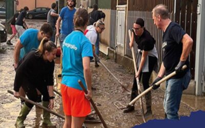 Group of volunteers shoveling mud from a flooded street, wearing gloves and casual clothes; muddy ground and water puddles visible.