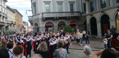 A street parade with a marching band in black-and-red uniforms playing brass instruments, while spectators line a European-style street with arches and a Foot Locker storefront nearby.