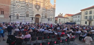 Large crowd seated for an outdoor concert in front of a striped stone cathedral.