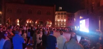 Nighttime outdoor concert or performance with a large crowd facing a bright stage on the right and historic arches on the left.