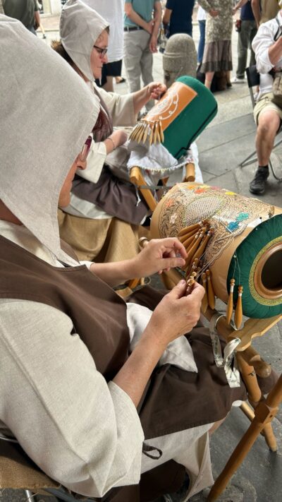 Two people in beige medieval-style garments weave decorative cords around large carved gourds, with onlookers in a plaza background.