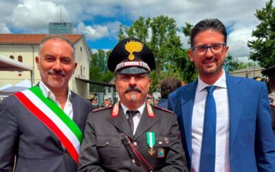 Three men posing at an outdoor ceremony: left in a suit with an Italian tricolor sash, center in military dress uniform with medals and hat, right in a blue suit and glasses.