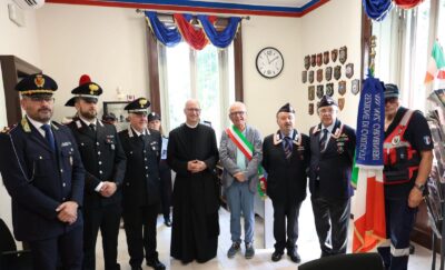 Group of uniformed officials and a priest posing in a ceremony room, shields on the wall and Italian flags nearby.