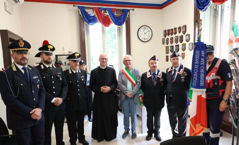 Group of uniformed officials and a priest posing in a ceremony room, shields on the wall and Italian flags nearby.
