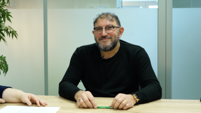 Smiling man with glasses and beard sits at a wooden table with papers, holding a green pen in front; office with frosted glass panels in the background.