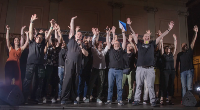 Group of people on a stage with arms raised in celebration, smiling at a concert or event moment.